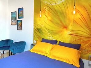 A bedroom with a bed, two chairs, a wall with framed pictures, and a large yellow floral tapestry. - Studio Moderne au Cœur de Rochefort - Vivez L'élégance et le Confort (Rochefort)