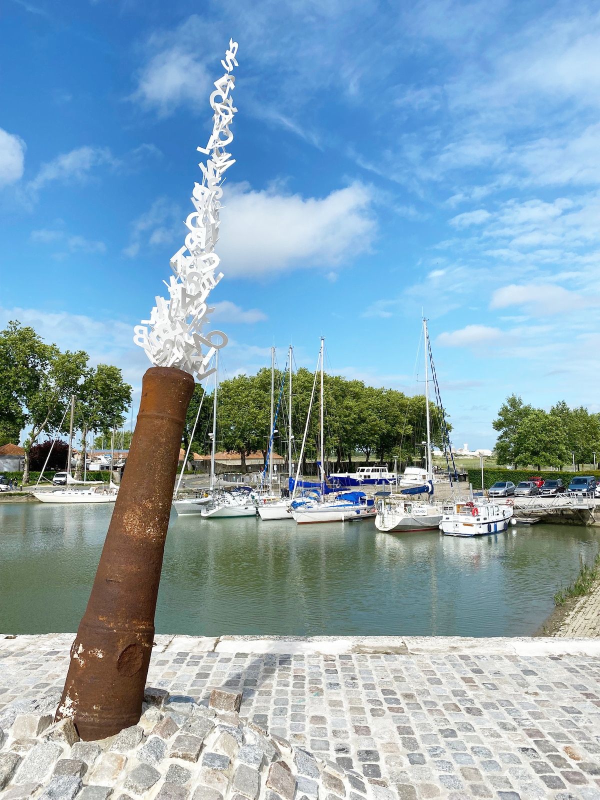 Une vue sur le front de mer, avec une sculpture en métal rouillé, des bateaux amarrés et une promenade pavée.