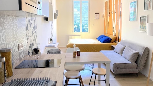 Una habitación de hotel moderna con cama, sofá, mesa de comedor con taburetes, zona de cocina y un amplio ventanal que proporciona luz natural.