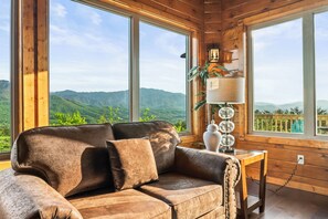 A wooden cabin with a large window offering a mountain view. Inside, there's a brown leather sofa with cushions. - Breathtaking Views - 10BR/Wedding & Reunion/Firepit/Space for 46/No Event Fee (Gatlinburg)