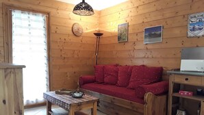 Une pièce chaleureuse avec des murs en bois, un canapé rouge, une table en bois recouverte d’une nappe à carreaux, un lampadaire et une horloge.