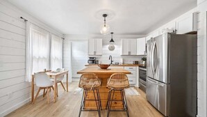Une cuisine moderne avec des armoires blanches, un réfrigérateur en acier inoxydable, un îlot central en bois avec deux tabourets, et un luminaire suspendu.