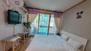 A hotel room with a bed, a TV, a desk with a chair, a potted plant, and a view of a swimming pool.