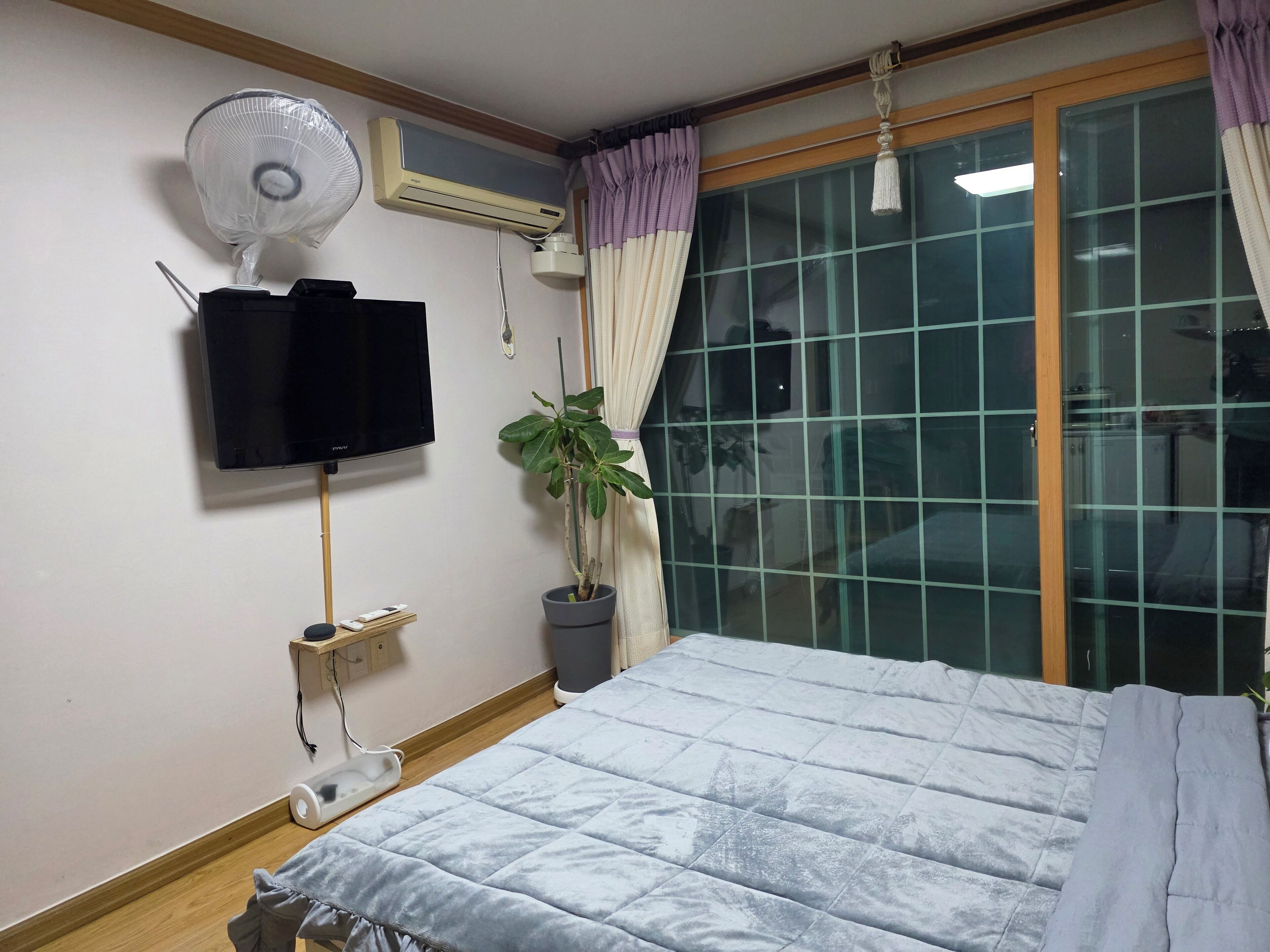 Ein Schlafzimmer mit einem Bett, einer Wand-Klimaanlage, einem Standventilator, einem Fernseher, einer Topfpflanze und einem Fenster mit Vorhängen.