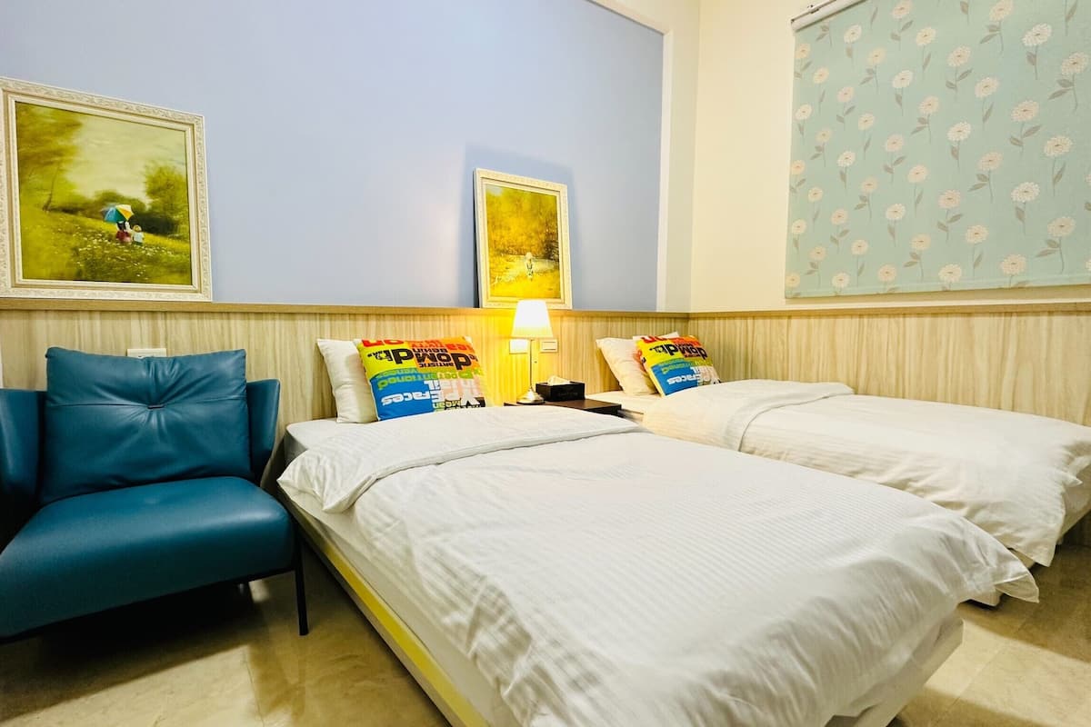 A hotel room with two beds, a blue chair, and two paintings on the wall.