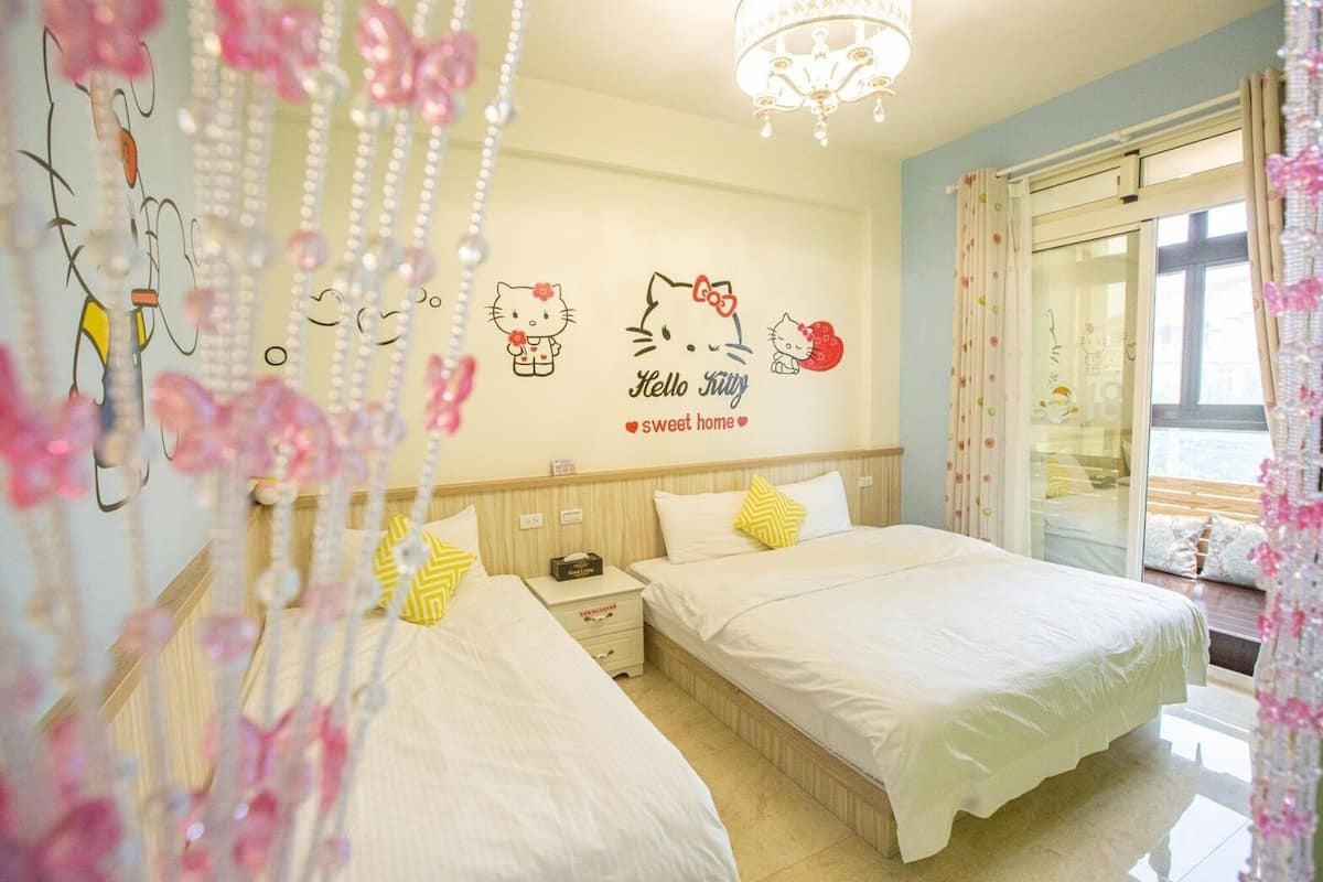 A bedroom with two beds, a wall decal with Hello Kitty, a chandelier, and a window with curtains.
