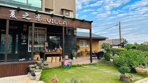 Una villa con fachada de madera, un patio con mobiliario exterior y un jardín con diversas plantas.