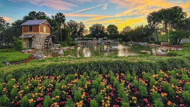 A rustic stone building with a wooden roof near a pond, surrounded by lush greenery and colorful flowers. - St. John's, 3 bdrm, 2 bath, 2022 built home. 10% off military, breast cancer (The Villages)