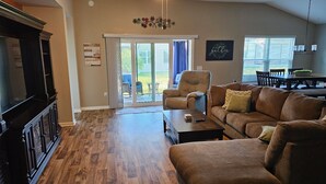 A spacious living room with a dark wood floor, a brown leather sofa, a coffee table, and a television on a cabinet. - St. John's, 3 bdrm, 2 bath, 2022 built home. 10% off military, breast cancer (The Villages)