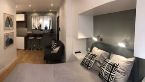 A modern bedroom with a large bed, a desk, and a kitchenette in the background. - Studio Moderne Proche Champs Elysée (Paris)