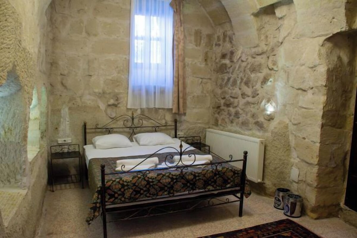 Een slaapkamer met stenen muren, een bed, een nachtkastje, een radiator en een raam met gordijnen.