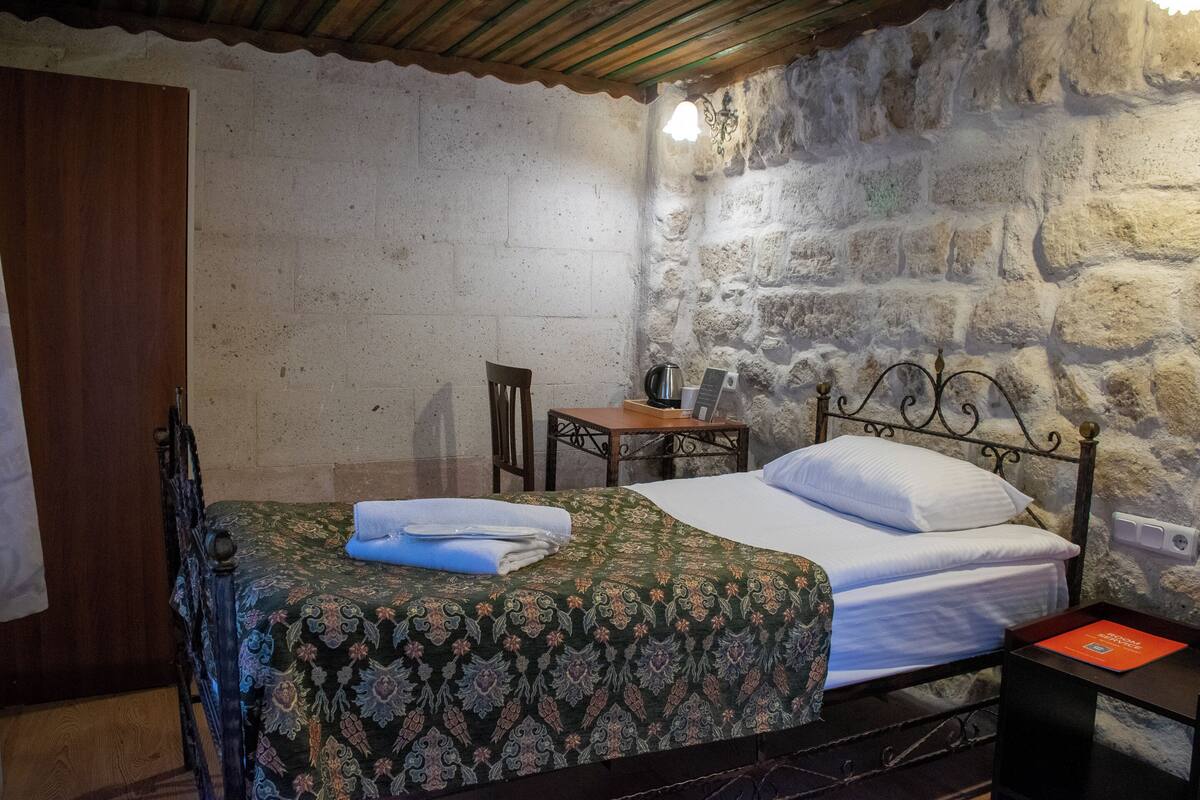 A single bed with a patterned bedspread, a wooden chair, a small table, and a stone wall.