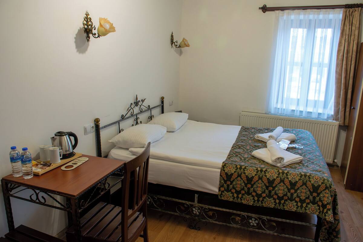 A neatly made bed with a floral bedspread, a bedside table with a kettle and water bottles, and a window with curtains.