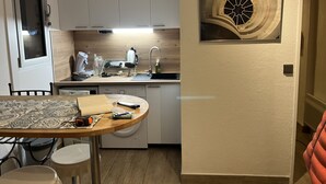 Ett kompakt kök med vita skåp, en inbyggd diskmaskin och en diskho. Det finns ett matbord med stolar och en dekorativ väggdekoration ovanför.
