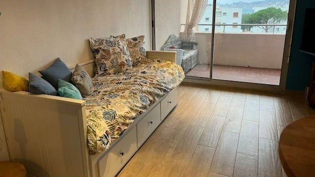 Ett rum med en säng, en soffa, en skjutdörr i glas som leder ut till en balkong och utsikt över byggnader och träd.