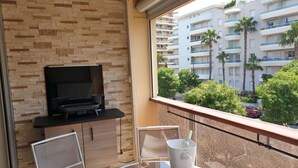 A balcony with a TV, a table with a wine glass and a bottle, and a view of a residential building with palm trees. - Beachside Studio with Terrace, A/C, Parking - Sleeps 4 in Fréjus (Fréjus)