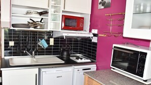 A compact kitchen with white cabinets, a black tiled backsplash, a red microwave, a white dishwasher, and a countertop with a kettle and a wine glass holder. - South-facing studio 4 pers, near center, terrace (Pralognan-la-Vanoise)