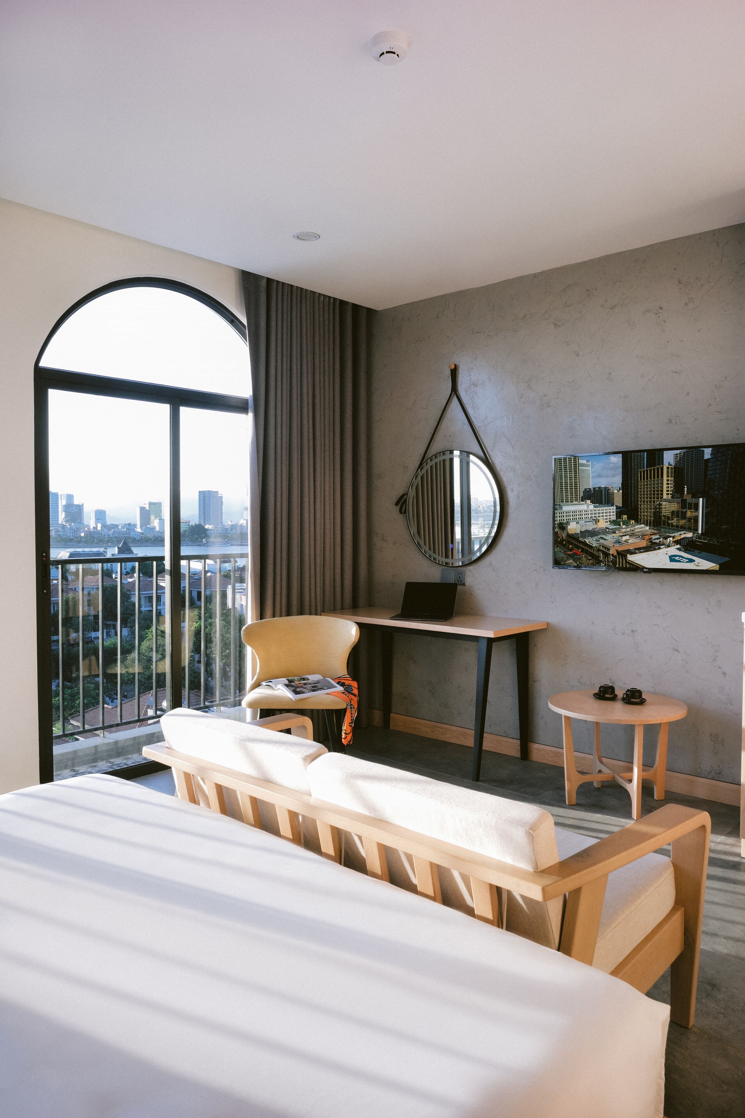 A hotel room with a large bed, a desk, a chair, a small table, a TV, and a view of the city.