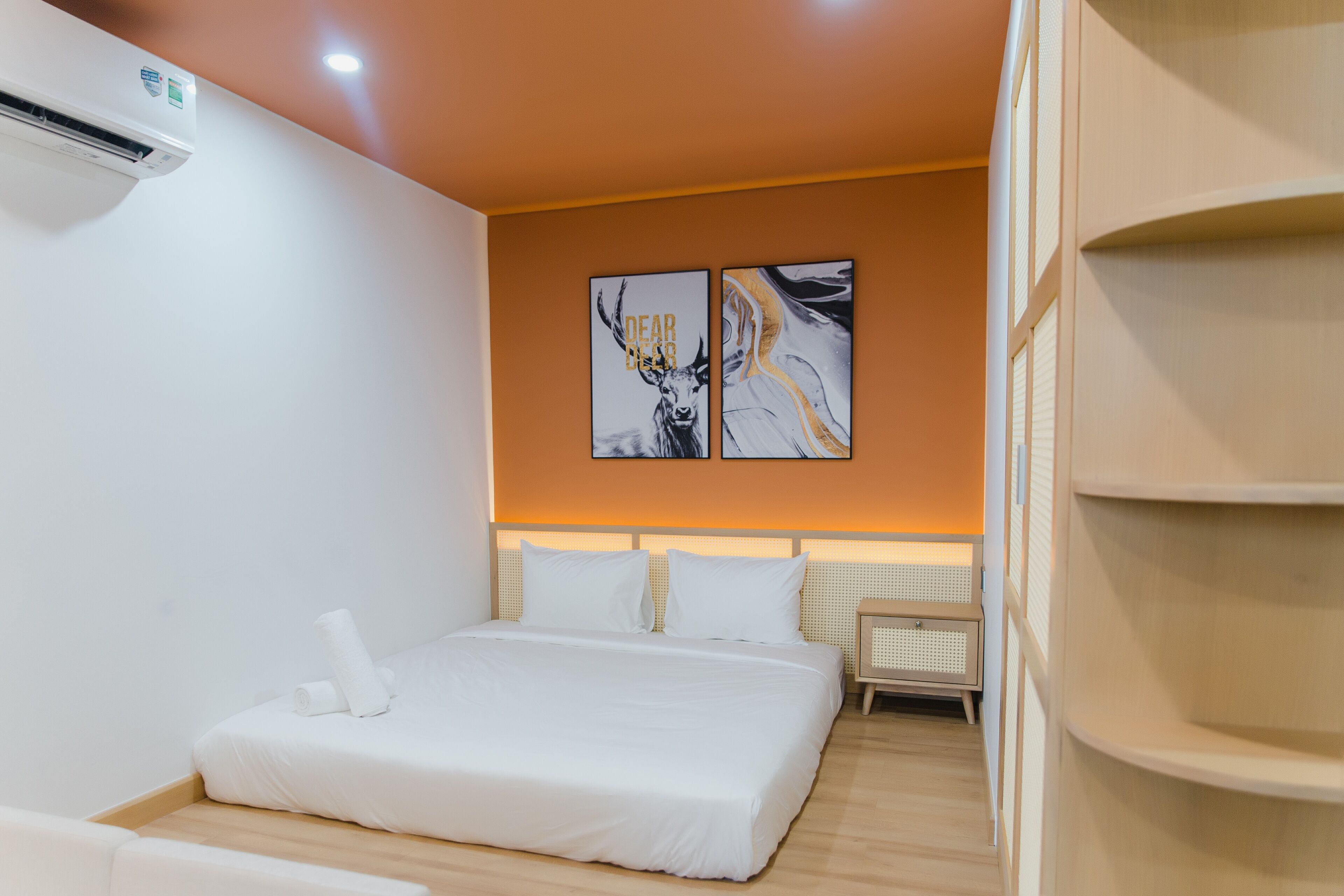 A hotel room with a bed, white bedding, a wall-mounted air conditioner, and two artwork pieces above the bed.