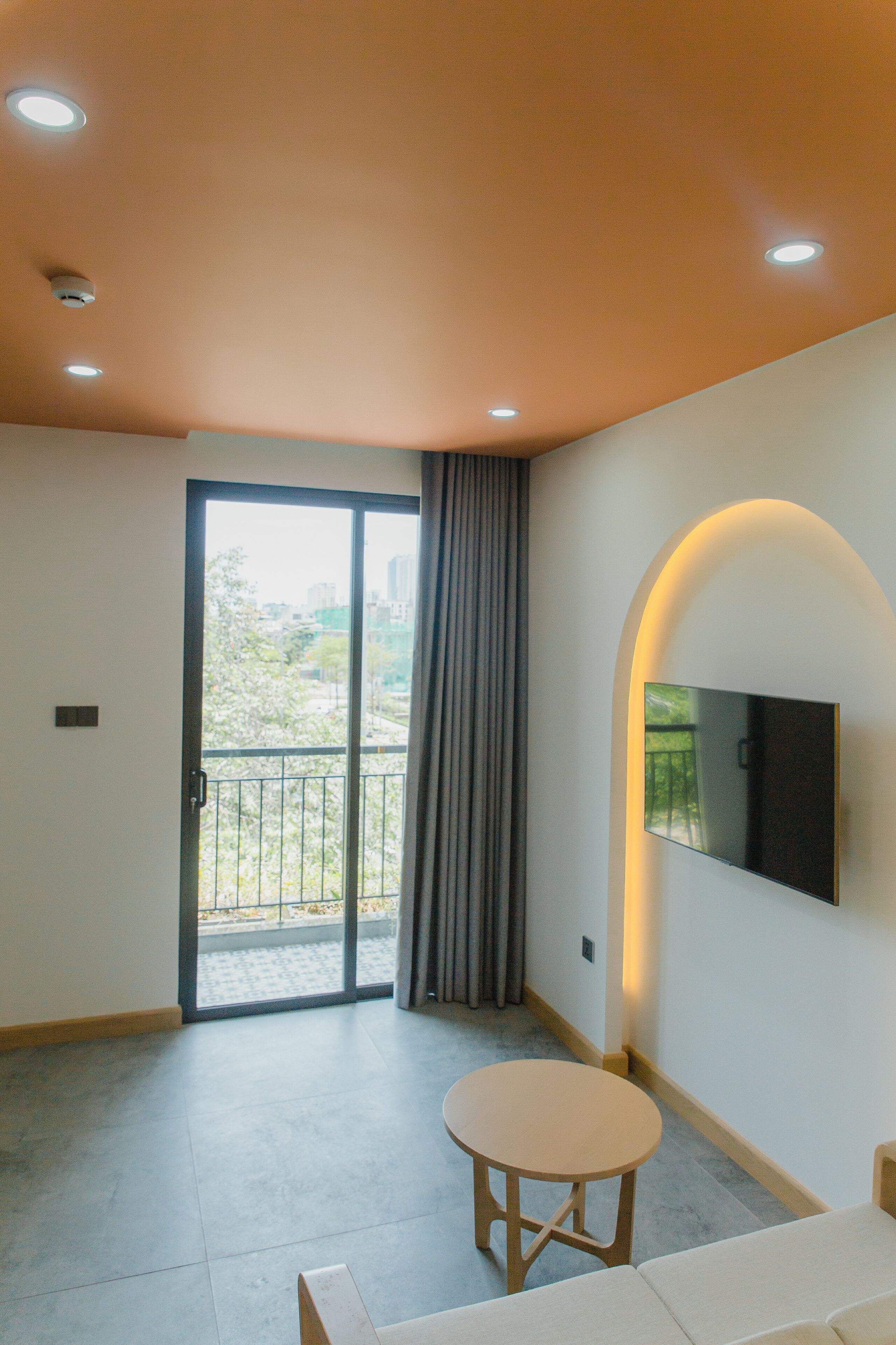 A modern living room with a sliding glass door leading to a balcony, a flat-screen TV mounted on the wall, and a round wooden table.