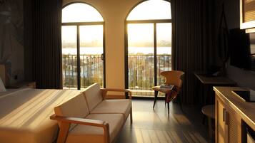 A hotel room with a bed, a sofa, a desk, and a TV. There are large windows with a view of buildings and a body of water.