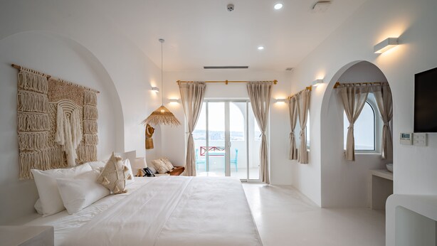A spacious bedroom with a large bed, a macrame wall hanging, and a view of the city through the balcony doors. - Happiness Hotel (Quy Nhon Dong)
