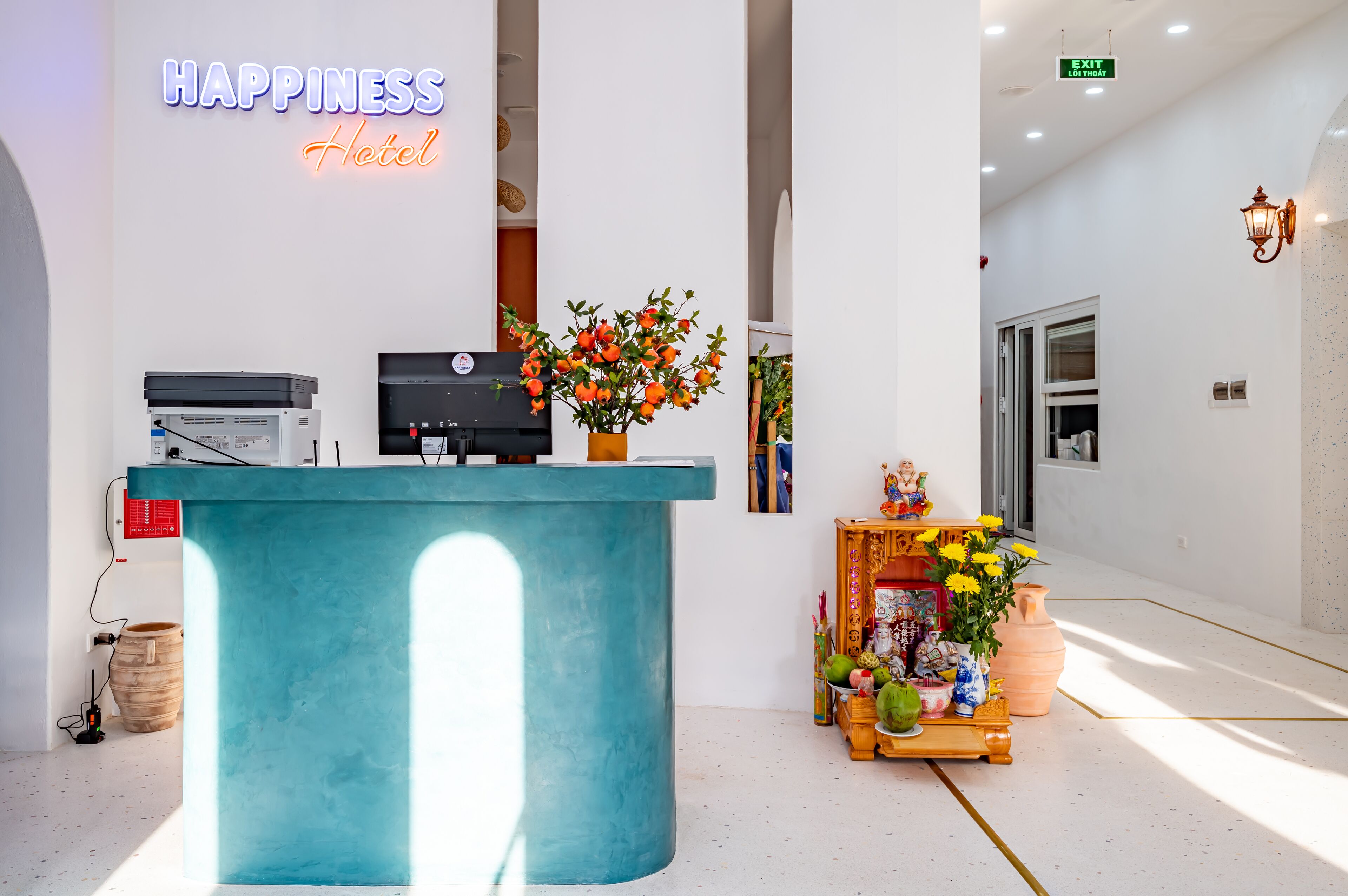 Vastaanottotiski, jossa on neonvalomainos, jossa lukee ”Happiness Hotel”, ruukussa oleva kasvi ja pieni alttari, jossa on tarjottuja esineitä.