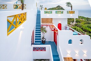 A modern hotel with a white exterior, blue steps, and red accents. It features a staircase leading to a balcony with a view of the sea and hills. - Happiness Hotel (Quy Nhon Dong)