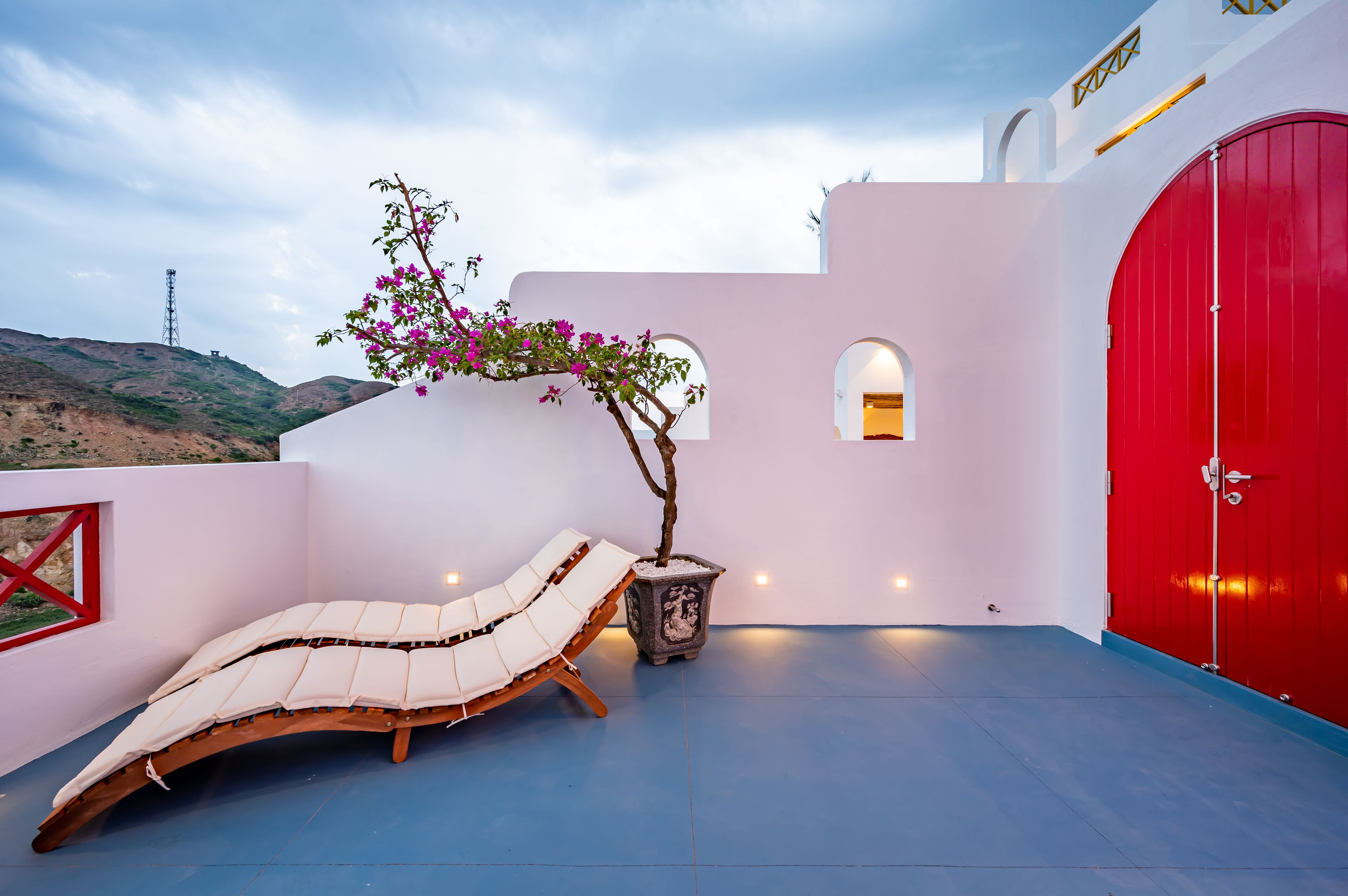 A sun loung chair, a potted plant, a red door, and a white wall with a small window.