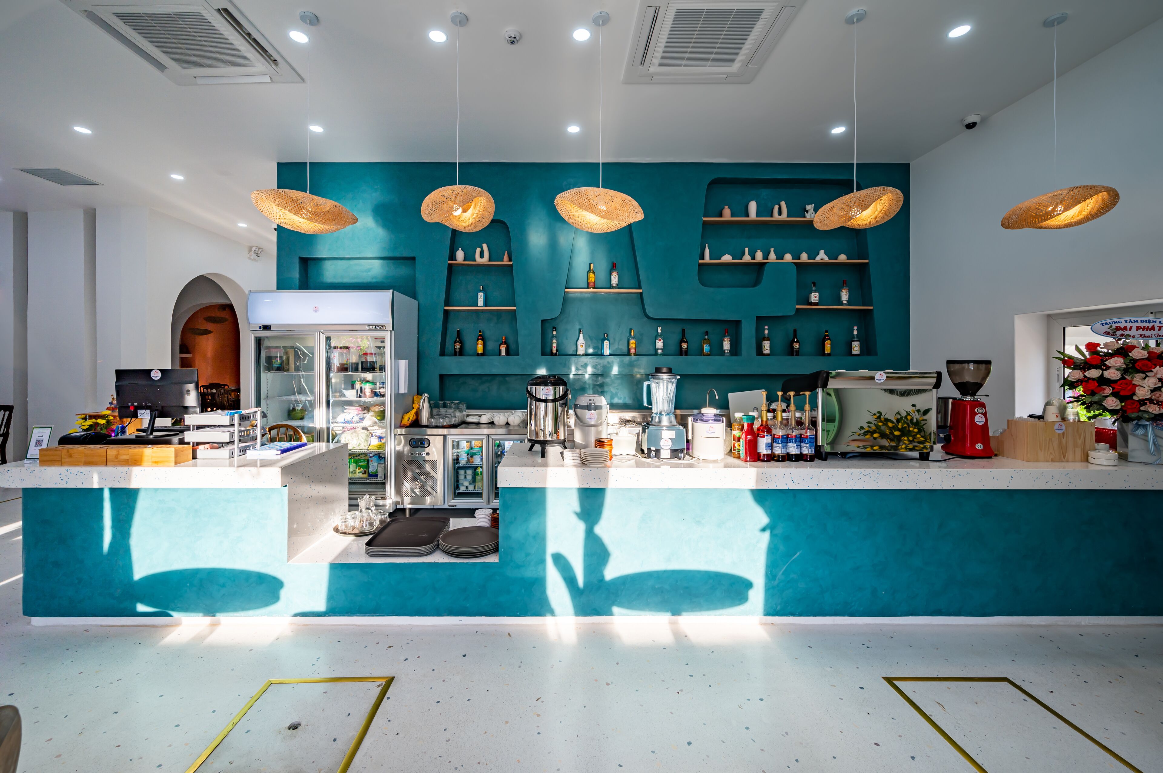 Eine moderne Bar mit Türkis-Farben, hängenden Korbschwingenlampen und einer Auswahl an Flaschen und Gläsern in Regalen.