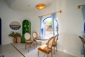 A dining area with a round table and wicker chairs, a green cabinet, and a window with blue curtains. - Happiness Hotel (Quy Nhon Dong)