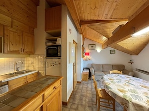 Een compacte keuken met houten kastjes, een magnetron en een koelkast. Er is een zithoek met een tafel en stoelen, en een bank.