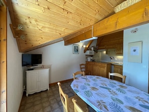 Een gezellige slaapkamer onder de dakpannen met houten balken, een televisie, een bed met een bedspread met tropisch dessin en een kitchenette met houten kastjes.