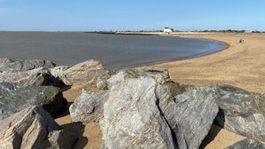 Une plage avec de gros rochers au premier plan, une étendue de sable et un petit bâtiment visible au loin.