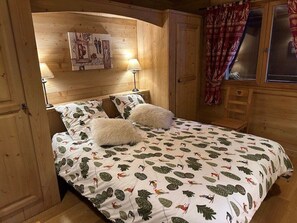 A wooden cabin bedroom with a bed, two white fluffy pillows, a nightstand with a lamp, a wooden wardrobe, and a window with curtains. - Comfortable chalet for 12 with WiFi and parking, near slopes (Crest-Voland)