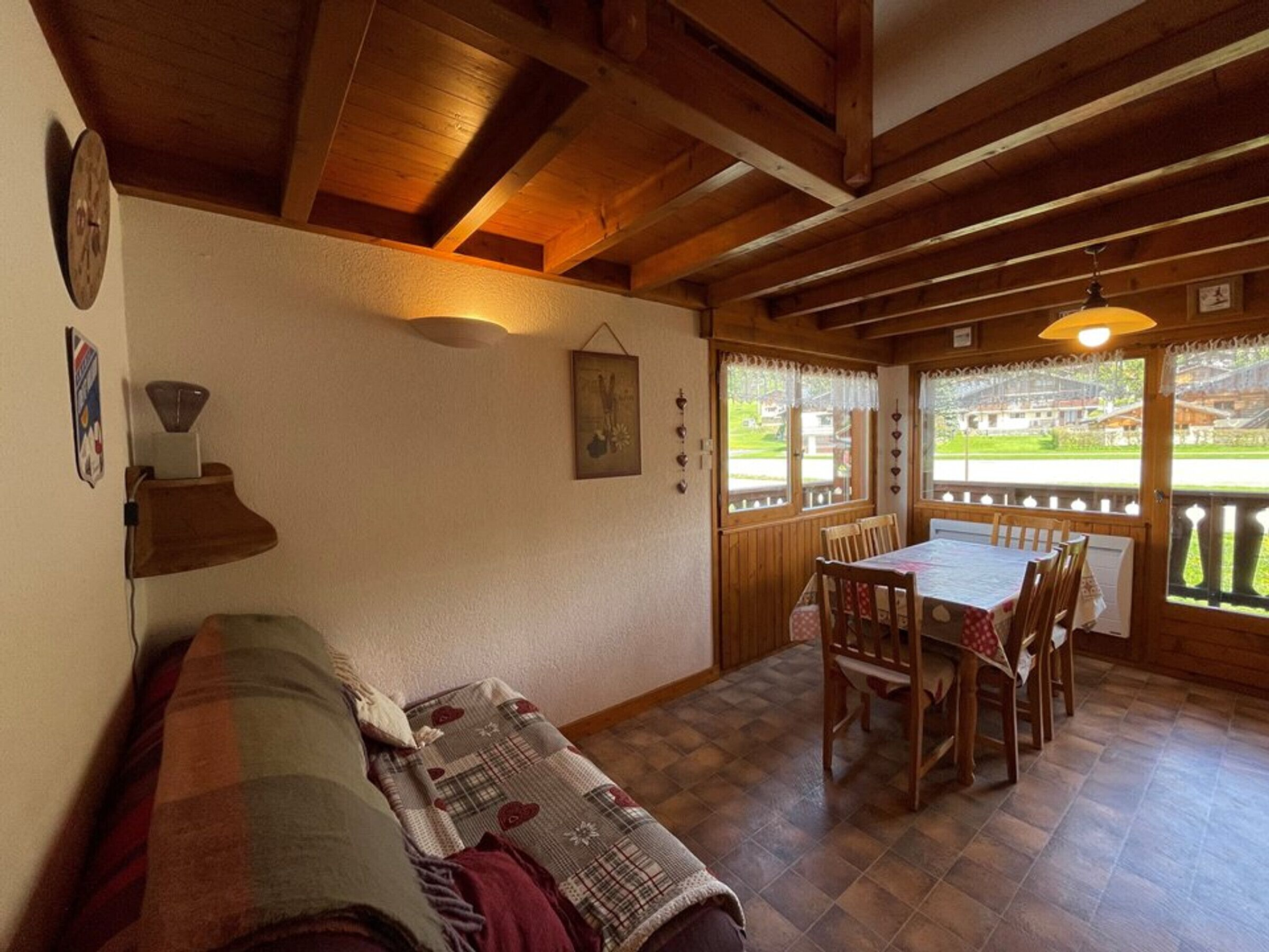 A cozy room with a wooden ceiling, a dining table with chairs, a sofa, and a window with a view of a green landscape.