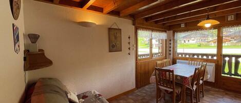 A cozy room with a wooden ceiling, a dining table with chairs, a sofa, and a window with a view of a green landscape.