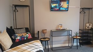 A bedroom with a bed, a TV, a small table, a shelf, and a hanging rack. - Apartment - Sleeps 5, Sun Terrace (Skegness)