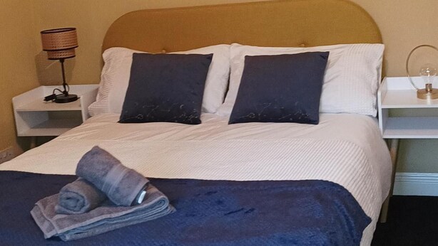 A neatly made bed with a white duvet and two dark pillows, a folded towel, and a lamp on a bedside table. - Apartment - Sleeps 5, Sun Terrace (Skegness)