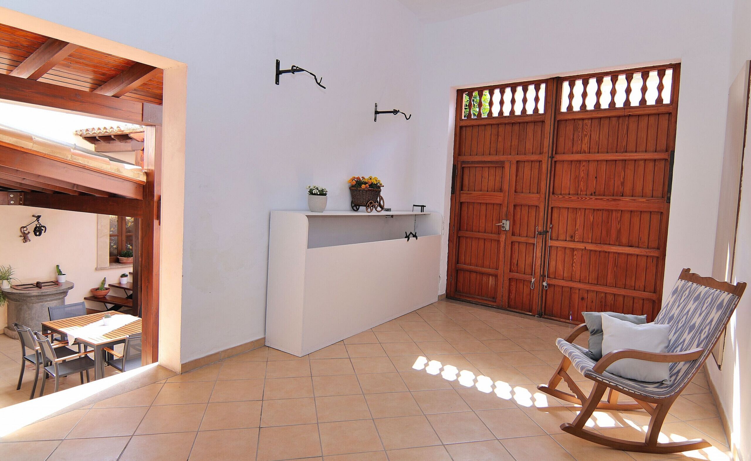 A patio with a wooden table, chairs, a rocking chair, a white counter with potted plants, and a large wooden door with a decorative panel.