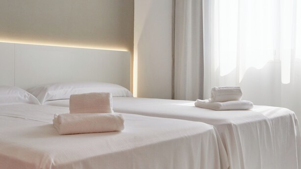 Habitación de hotel con dos camas, cada una con toallas ordenadas, una paleta de colores neutros y un ventanal con cortinas translúcidas.