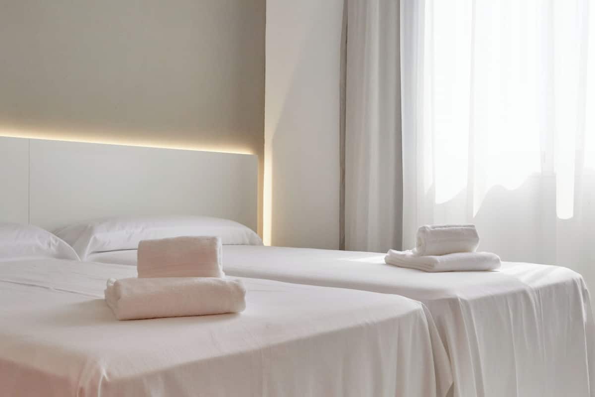 Habitación de hotel con dos camas, cada una con toallas ordenadas, una paleta de colores neutros y un ventanal con cortinas translúcidas.