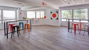 Una habitación amplia con techo alto, ventanas grandes y una zona central con taburetes de colores y un mural en la pared.