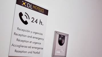Un letrero de hotel con aviso de recepción las 24 horas y servicios de emergencia, además de una cámara de seguridad montada en la pared.