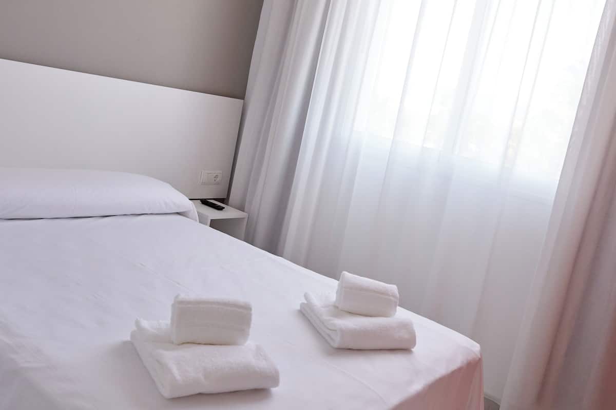 Una cama bien hecha con ropa blanca y toallas, una mesita de noche con lámpara, y cortinas translúcidas que dejan entrar la luz natural.