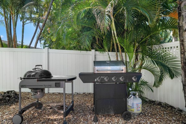 A portable Weber grill with a side table and a gas bottle, set against a backdrop of a white fence and tropical foliage.