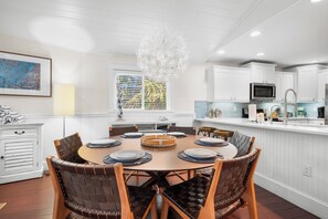 Cozinha moderna com armários brancos, sala de jantar com mesa redonda e cadeiras de fibra, e um lustre decorativo.