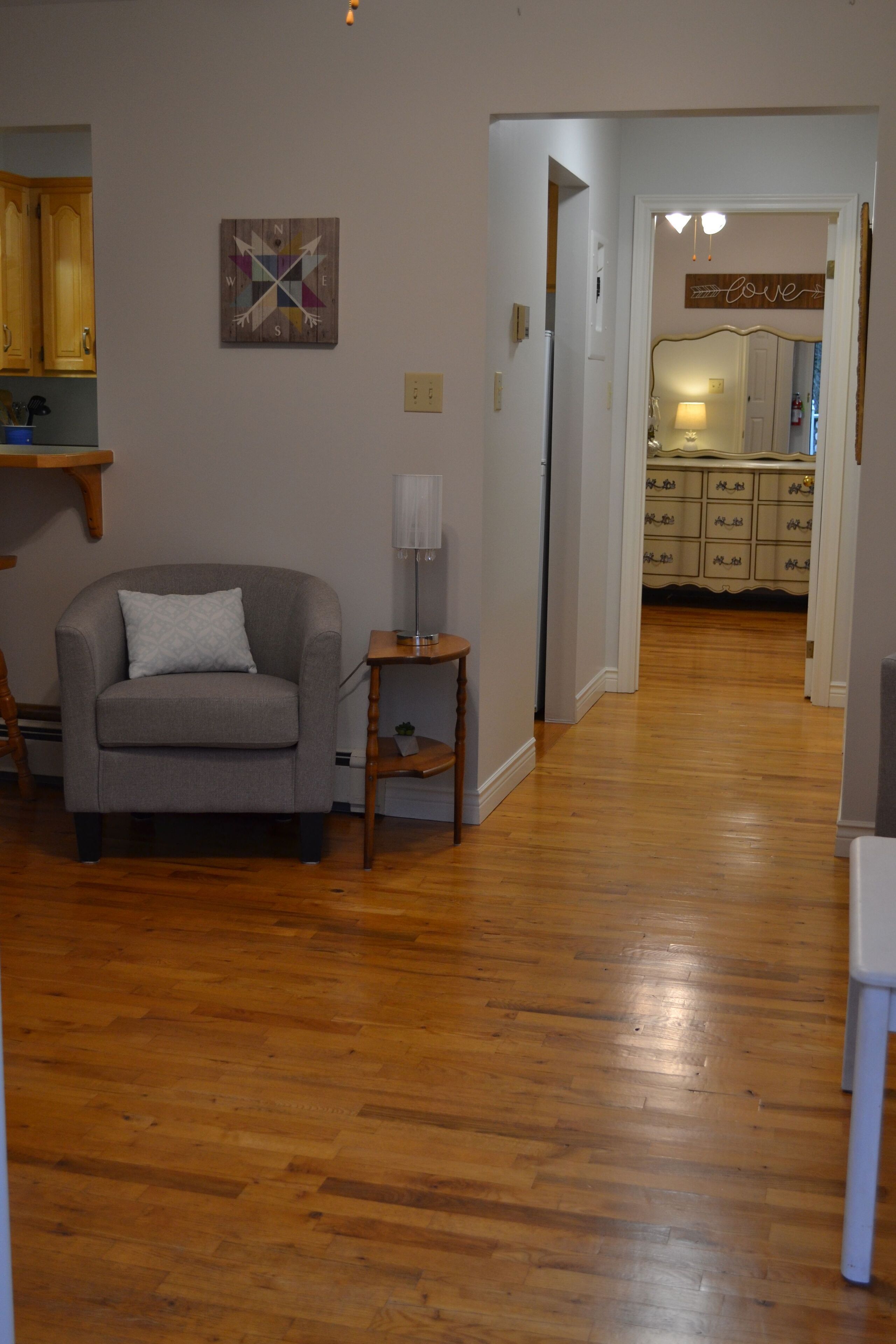 A living room with a grey sofa, a small table with a lamp, wooden flooring, and a kitchen visible through an open doorway.