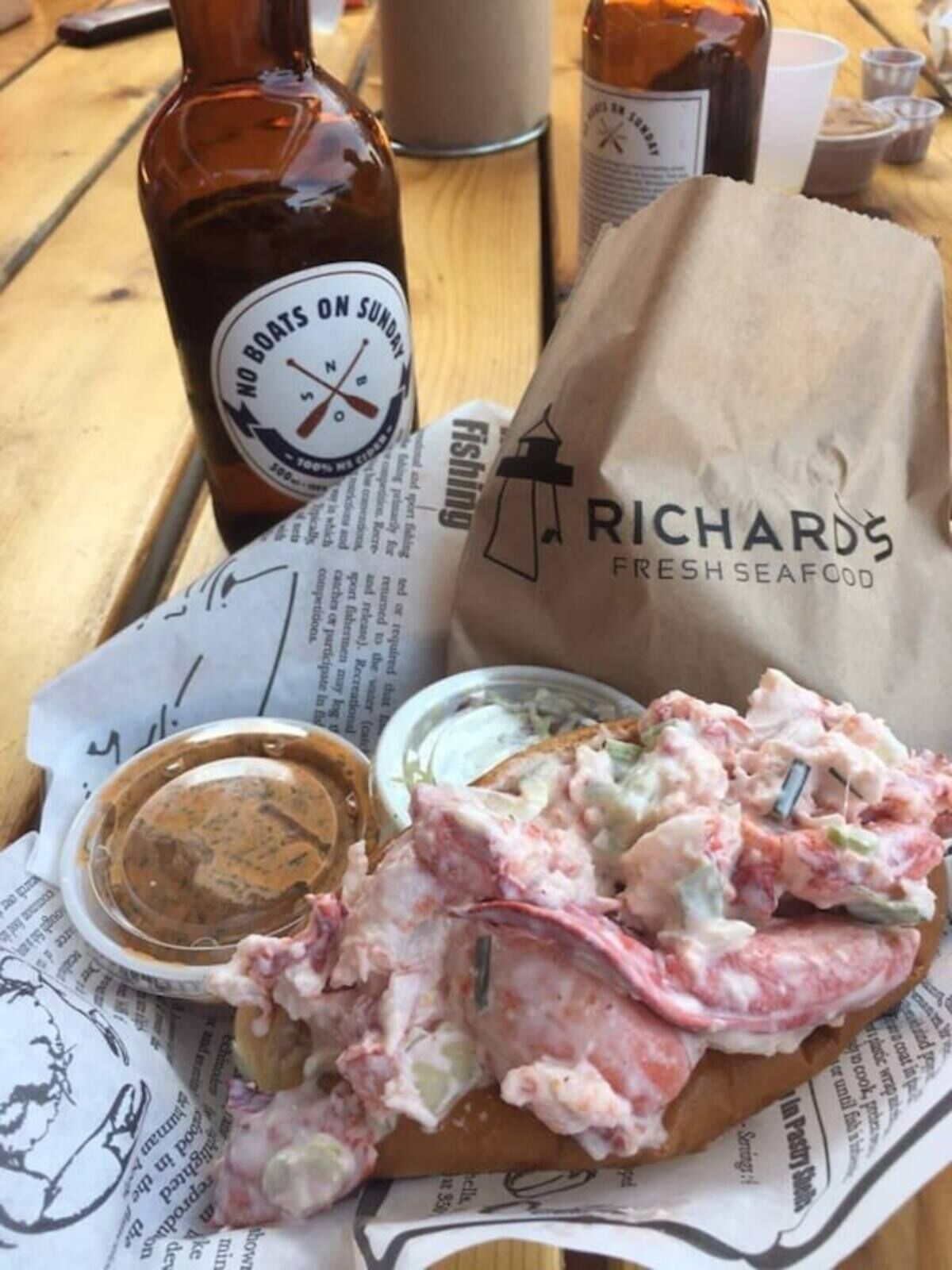 Ein Sandwich mit Soße, eine Bierflasche und eine Papiertüte mit Meeresfrüchte-Logo auf einem Holztisch.