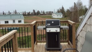 Una terraza de madera con una parrilla Weber, con vistas a una zona verde con casas y árboles.